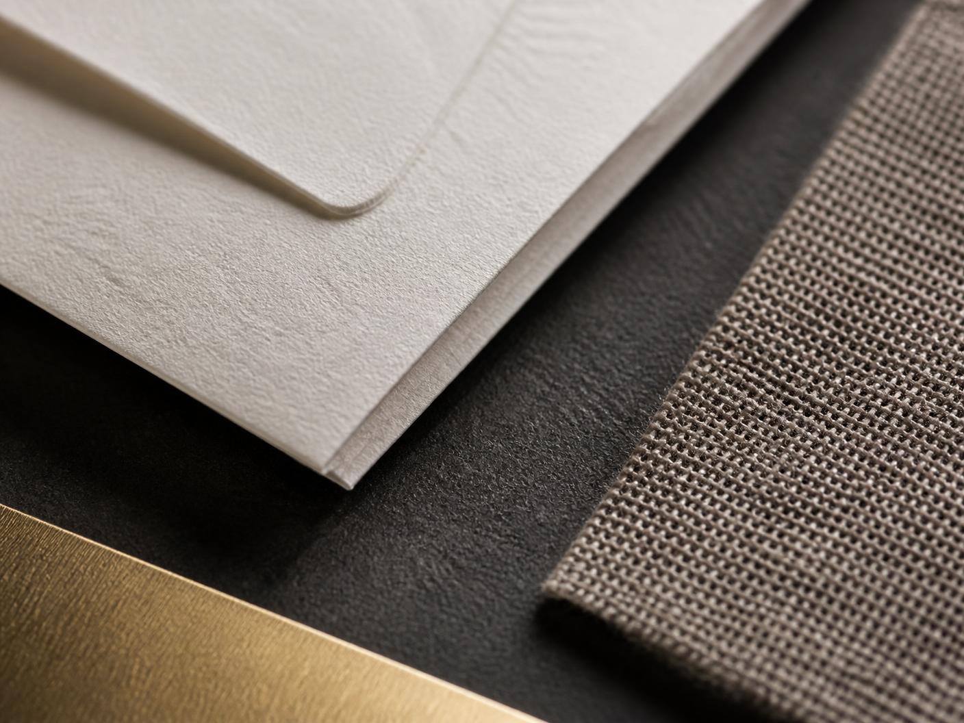 Close detail of an ivory letterpress paper edge with a blind deboss, a taupe woven linen swatch and a brushed gold trim resting on a charcoal surface